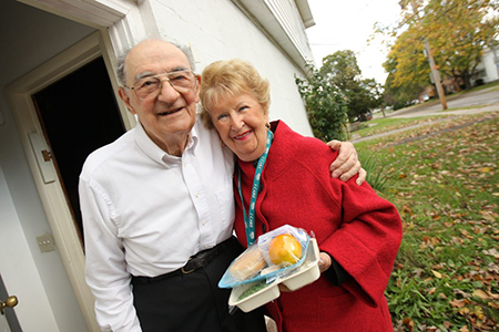 Woman delivering food to an older man outside the front door of his home.