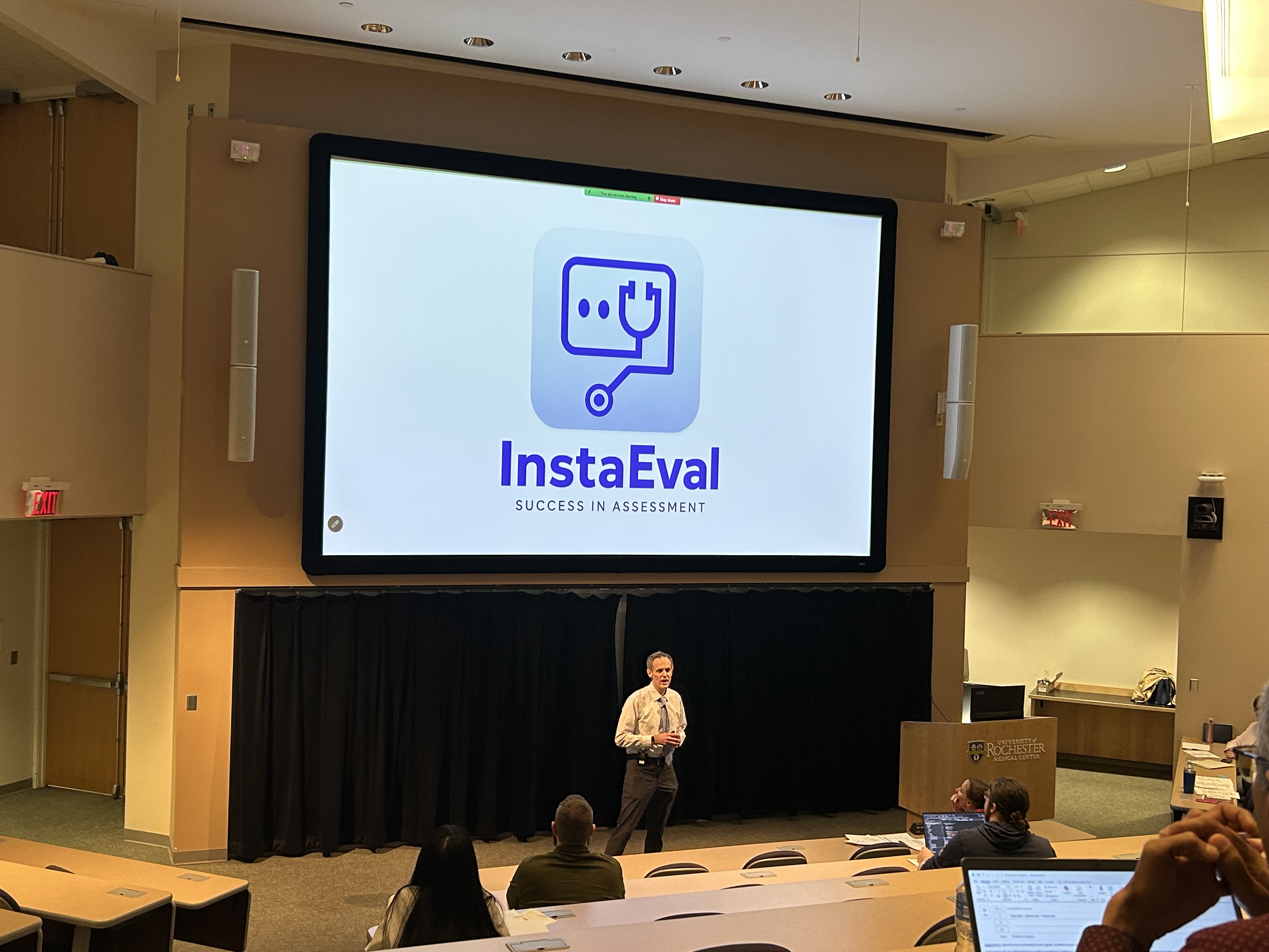 Dr. Stone presenting to an audience, with the InstaEval logo behind him on a large screen