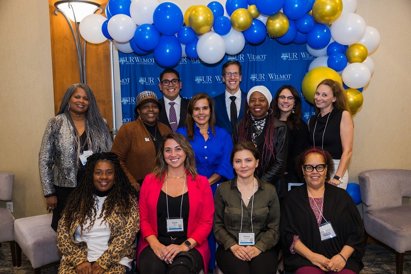A group of staff and community partners stand and smile for a photo in front of blue, gold and white balloons and a Wilmot Cancer Institute backdrop.