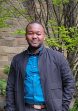 person standing outside in front of brick wall and tree, wearing a blue button up and navy jacket