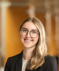 Person with long hair wearing glasses and blazer smiling at camera