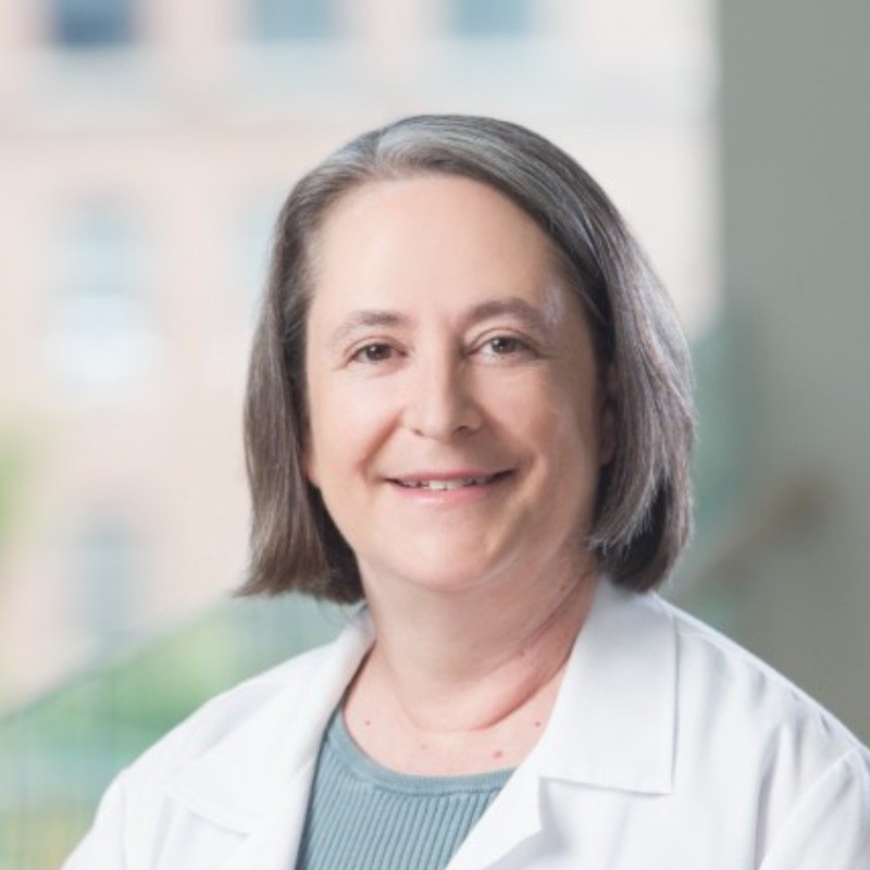 Dr. Eva Pressman, a smiling woman with straight gray hair, wearing a white lab coat over a light blue top, stands indoors with a blurred background.