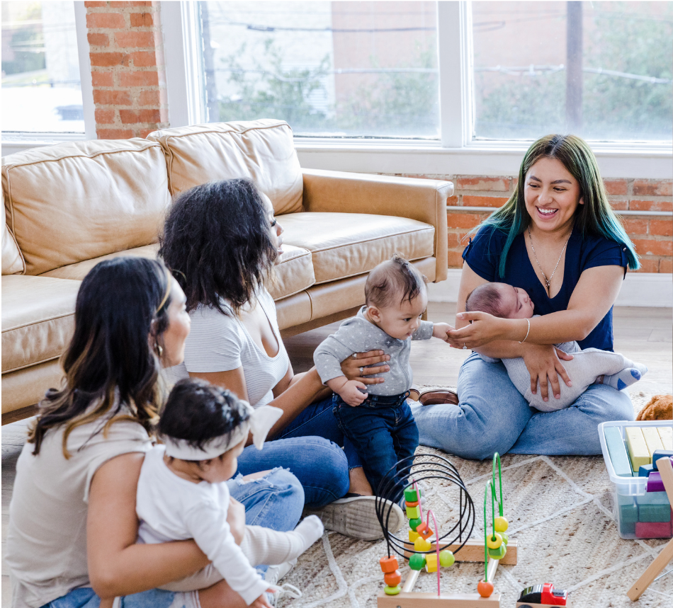 Group of three moms holding infants playing on the floor inside with a couch nearby