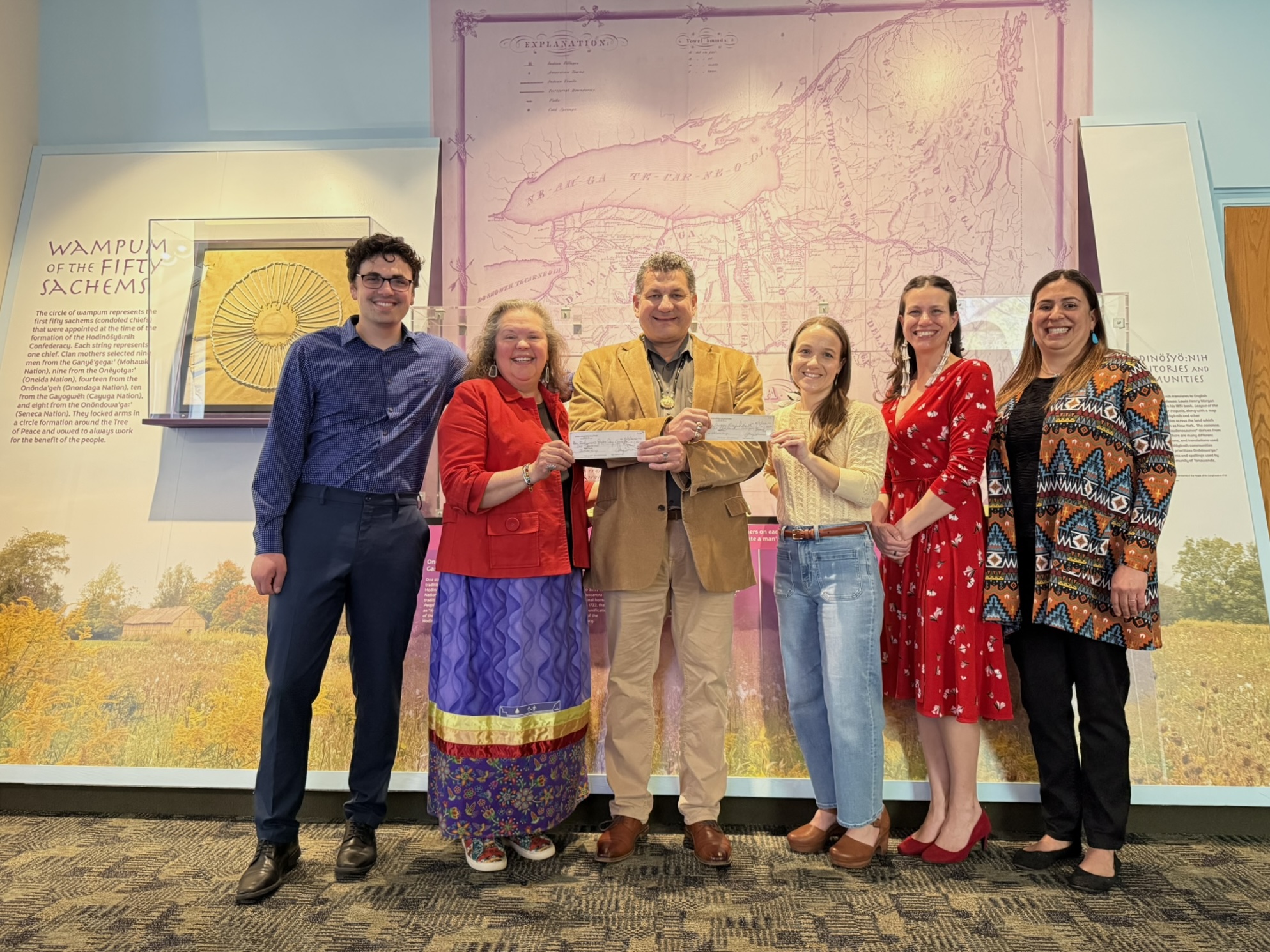 six individuals stand in front of wall with a map of New York State and Indigenous education, with the person in the middle handing each person next to him a paper check
