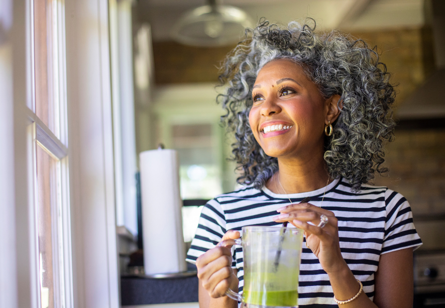 woman standing near window in home setting wearing a striped shirt and holding a green smoothie drink