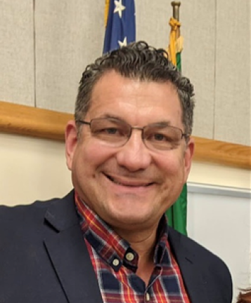 person wearing glasses and blazer with plaid button down shirt smiles at camera and stands in front of flags