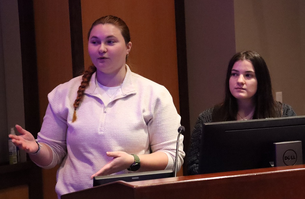 Rymal and Smith, female students, presenting in front of podium and computer