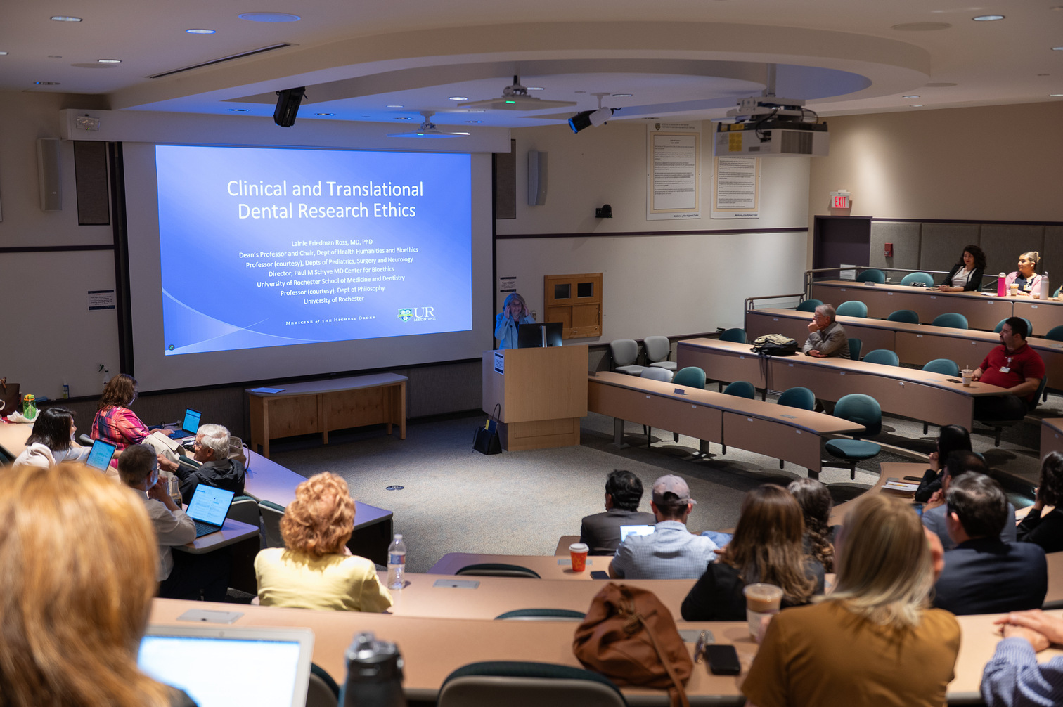 An oral presentation during the 2025 AADOCR conference, in a large lecture hall with rows of curved tables facing a large powerpoint screen in the center of the room.