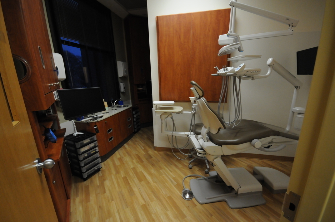 A roughly triangular patient treatment room with a window, featuring a dental chair and 2 monitors.