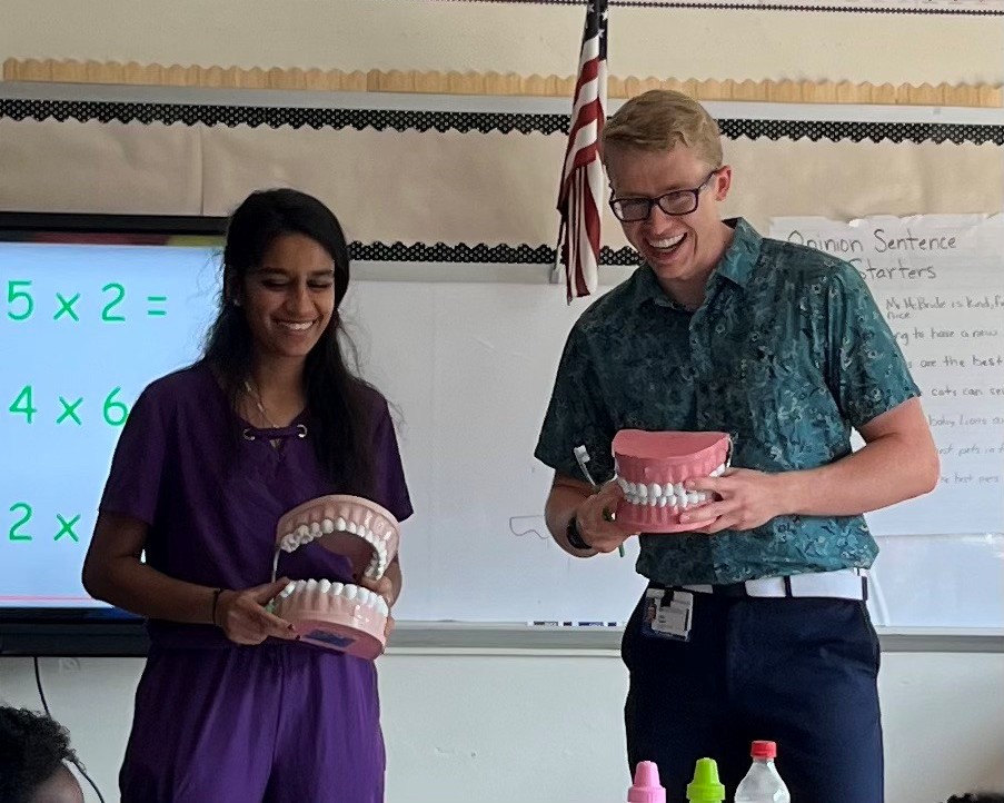 Two people stand at the front of a classroom holding oversized models of teeth. One person also holds a toothbrush and is energetically talking to the classroom.