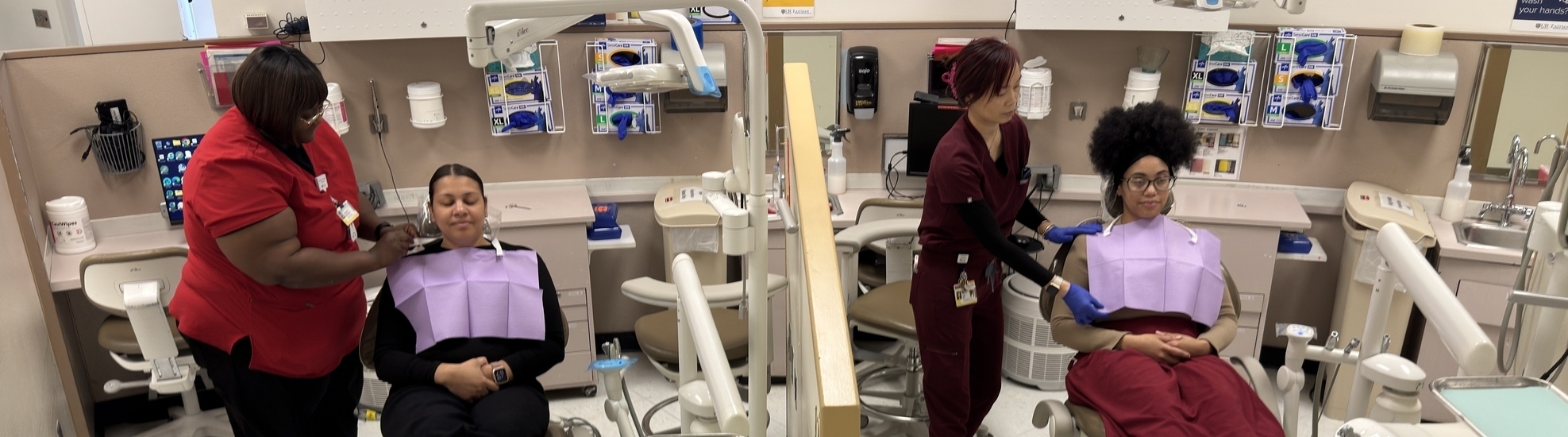 Two dental assistants work side-by-side getting patients comfortable in dental chairs.