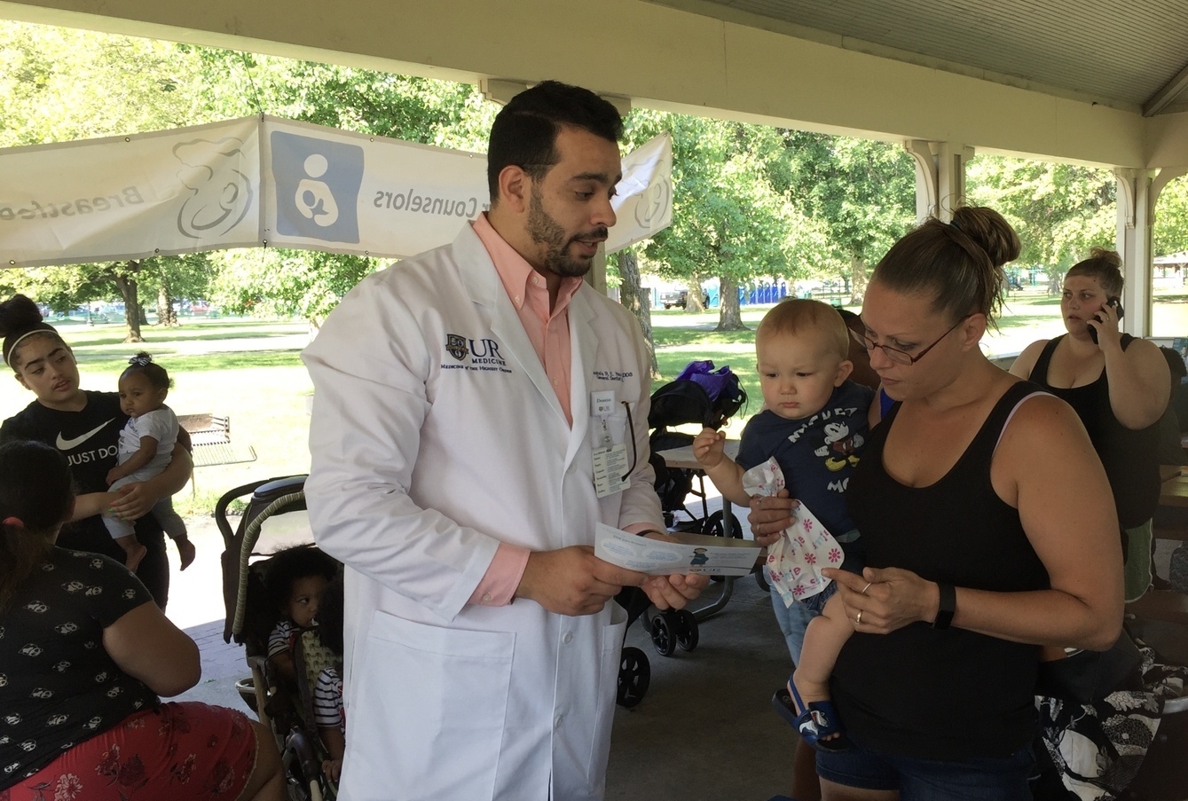 A dentist in a lab coat explains a piece of paper to a woman holding a baby. Other women holding babies can be seen in the background.