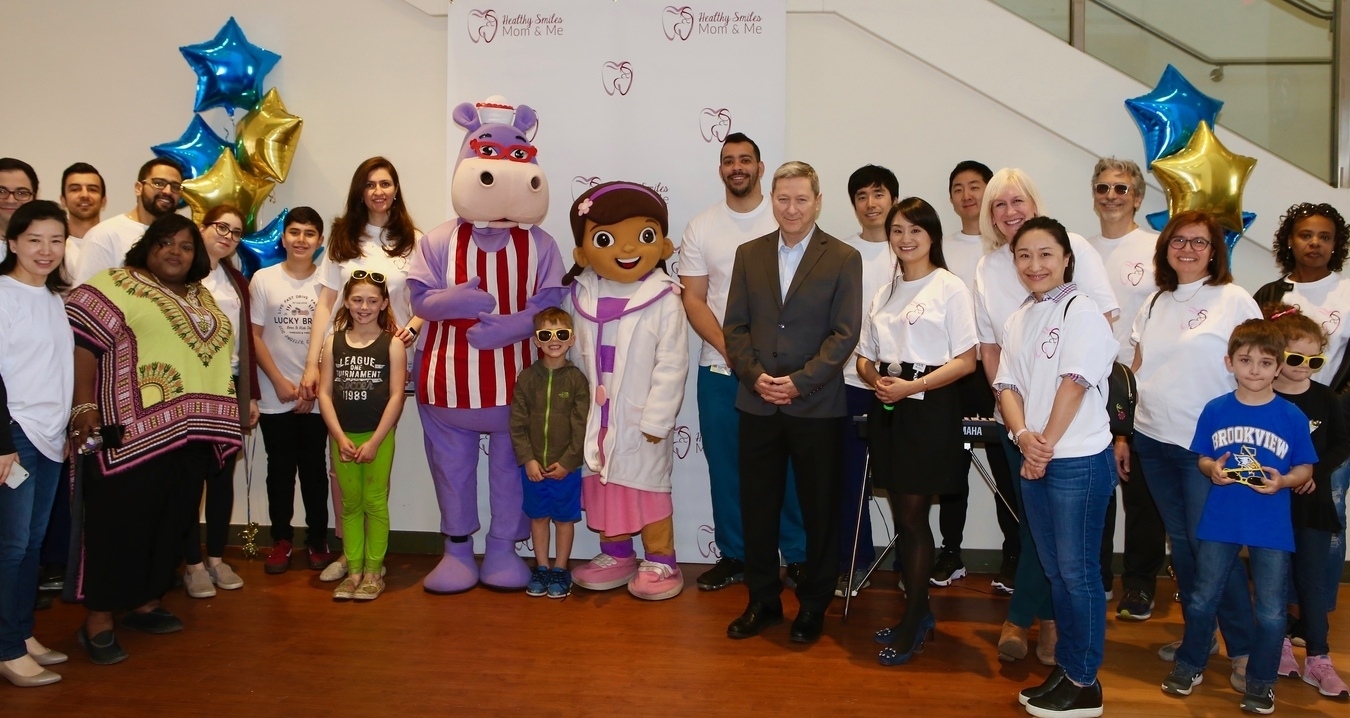 Multiple dentists and patients pose with two people dressed in mascot costumes of a purple hippo and a female dentist with an oversized plush head.
