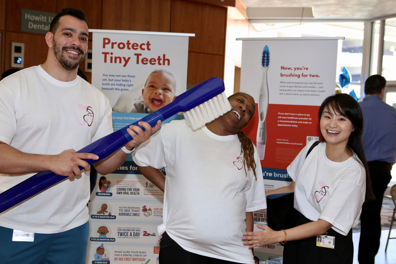 Three dentists laugh in front of a banner that says "Protect Tiny Teeth". One of the people is jokingly holding up a giant toothbrush to the face of the person in the middle.