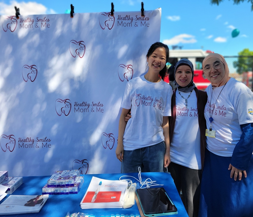 Three dentists wear matching event t-shirts and stand next to an informational table at an outdoor event.