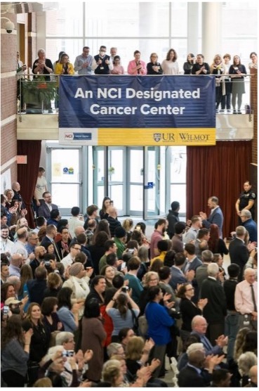 A blue and yellow banner with the words "An NCI Designated Cancer Center" hangs from a railing on the second floor of the Flaum atrium at the University of Rochester School of Medicine and Dentistry, while a large crowd gathers below and behind