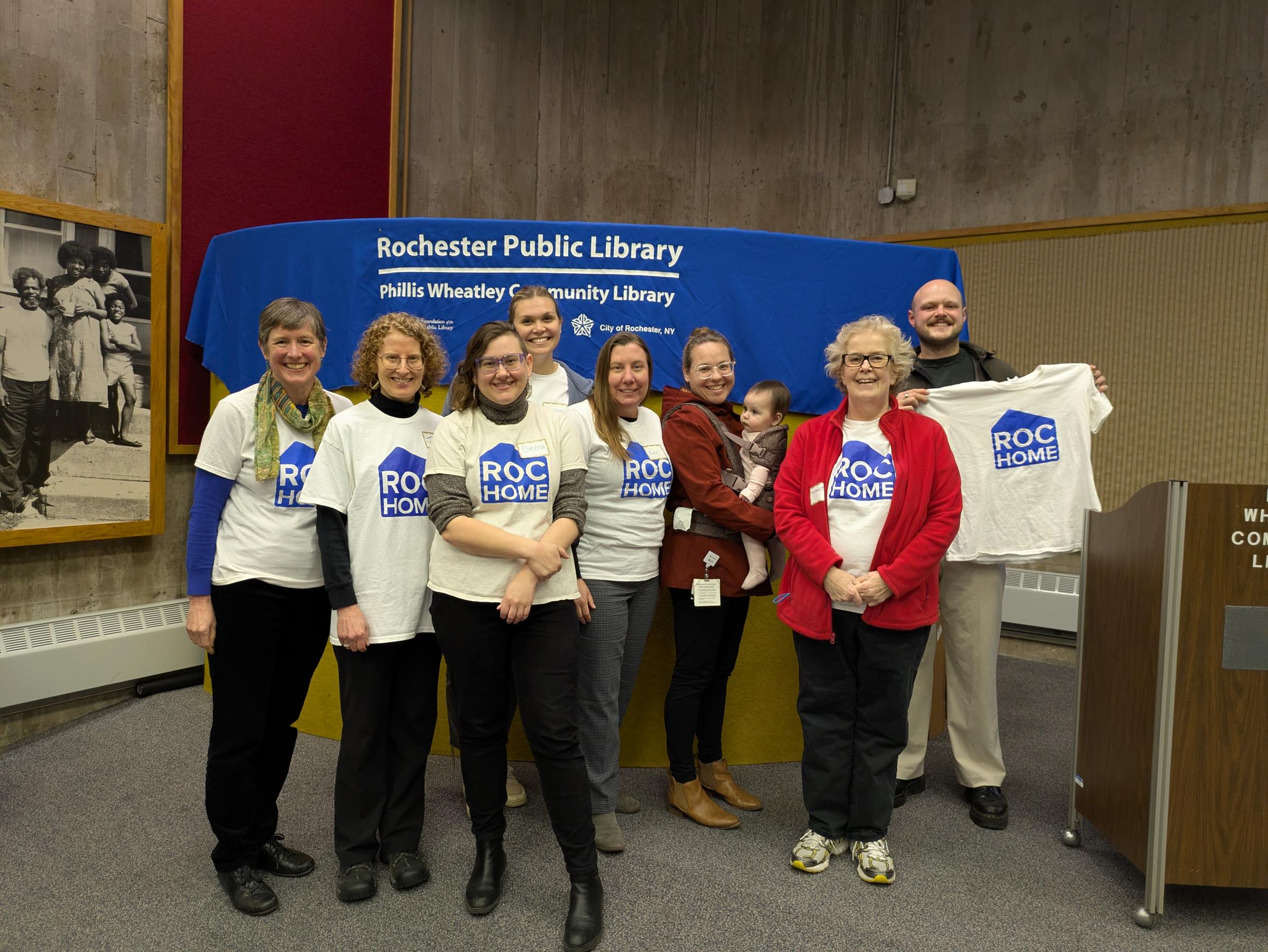 A group of people stands wearing matching white t-shirts with blue ROC Home logos on the front, in front of a Rochester Public Library sign, smiling and posing for a photo together.