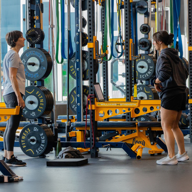 Woman working out in a gym, using weights and fitness equipment, talking with her trainer..