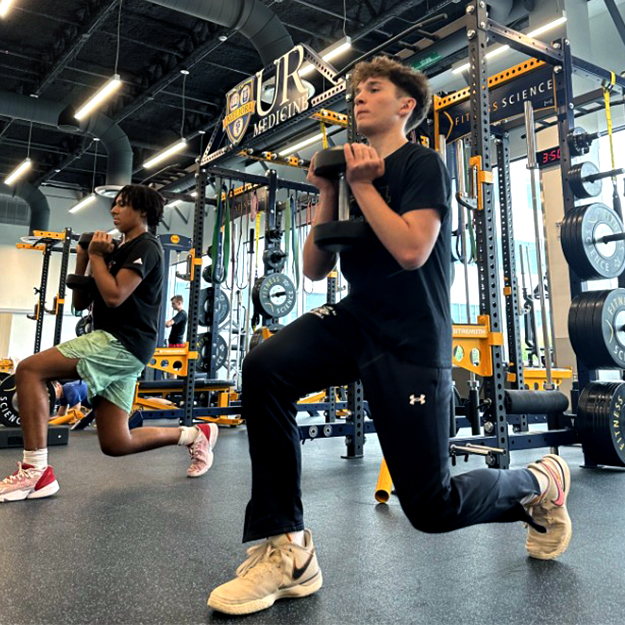 Two young men in a gym are doing squats, focusing on their form and strength training together.