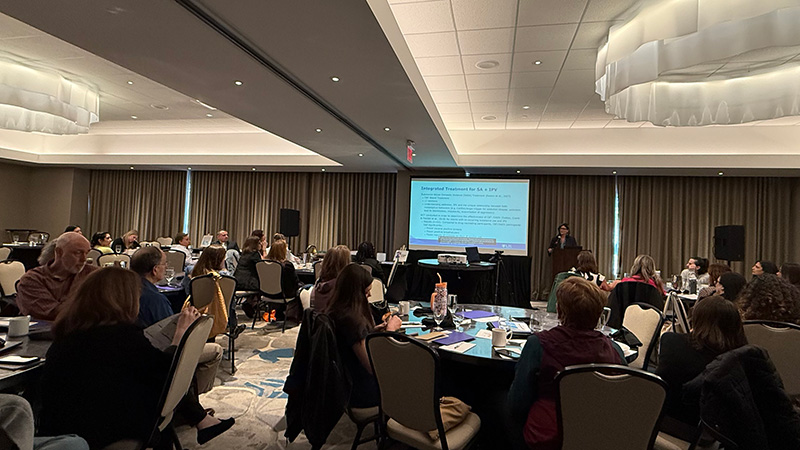 Attendees seated at tables watch a presenter speaking at a conference.