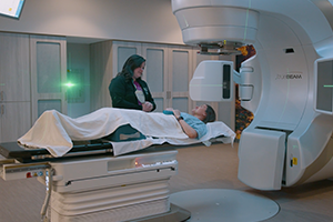 Patient receives radiotherapy on a treatment table beside a large medical linear accelerator, while a clinician stands nearby
