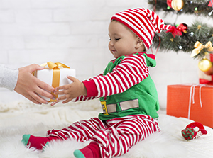A toddler dressed in a colorful elf outfit sits accepting with both hands a small gift box wrapped in gold ribbon.