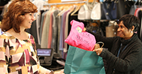 customer is seen picking up their order from gift shop staff, who is in the motion of placing a pink sweatshirt into a paper bag.