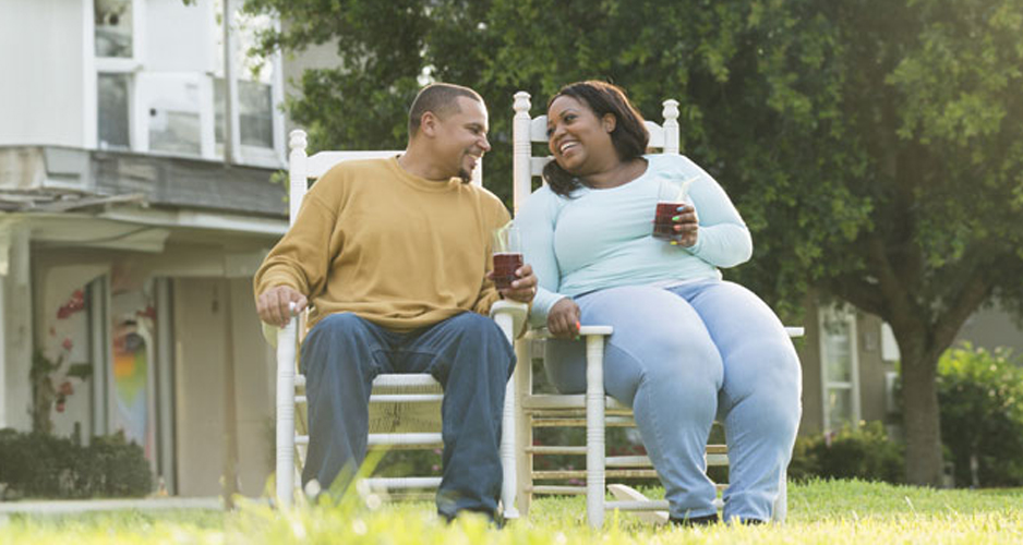 A man and woman sits side by side on a rocking chair, holding glasses of iced tea sharing a peaceful moment together in a warm atmosphere.