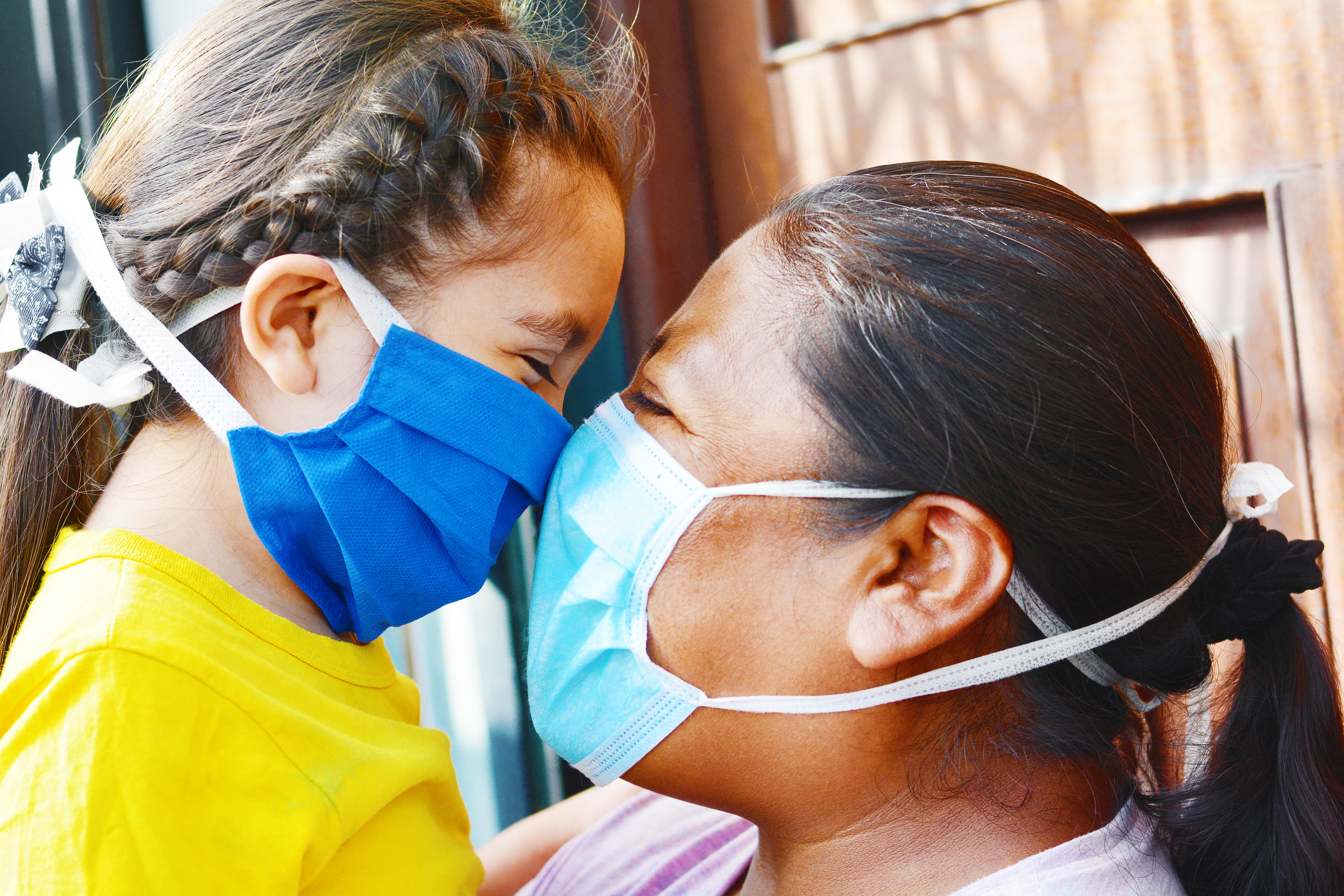 Native American woman and young girl, both wearing surgical masks, embrace and touch noses, smiling.